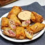 A white platter holding crispy reuben egg rolls. Some of the egg rolls are halved to show the cheesy inside filled with sauerkraut and diced corned beef. They are served with a wooden bowl of Russian dressing.