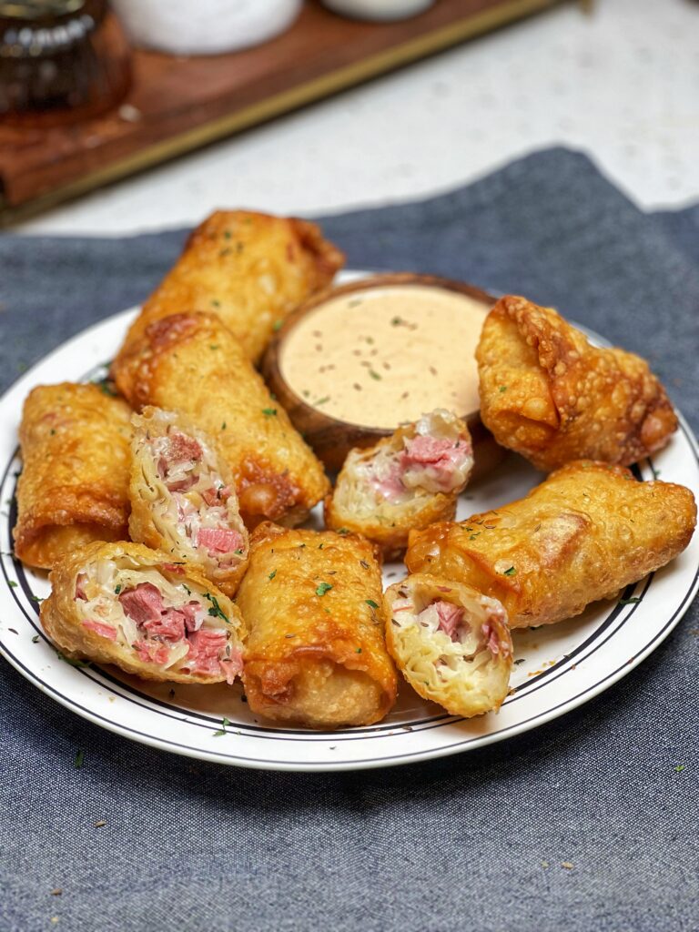 A white platter holding crispy reuben egg rolls. Some of the egg rolls are halved to show the cheesy inside filled with sauerkraut and diced corned beef. They are served with a wooden bowl of Russian dressing.