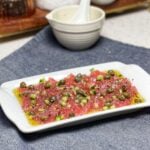 A small white rectangular platter with fresh tuna crudo. The raw tuna is sliced thin and topped with tiny diced cucumbers, red onion and drizzled with a lemon caper sauce.