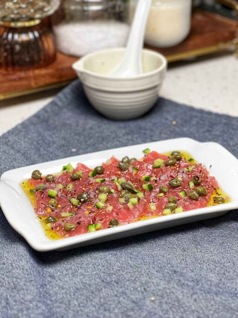 A small white rectangular platter with fresh tuna crudo. The raw tuna is sliced thin and topped with tiny diced cucumbers, red onion and drizzled with a lemon caper sauce.