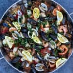 a pan of easy seafood paella filled with mussels, clams, lemon wedges and shrimp sitting on blue concrete