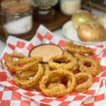 Crispy Vidalia onion rings sitting on red checkerboard parchment paper with a wooden bowl of dipping sauce.