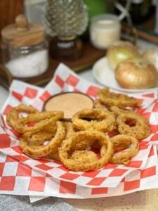Crispy Vidalia onion rings sitting on red checkerboard parchment paper with a wooden bowl of dipping sauce.