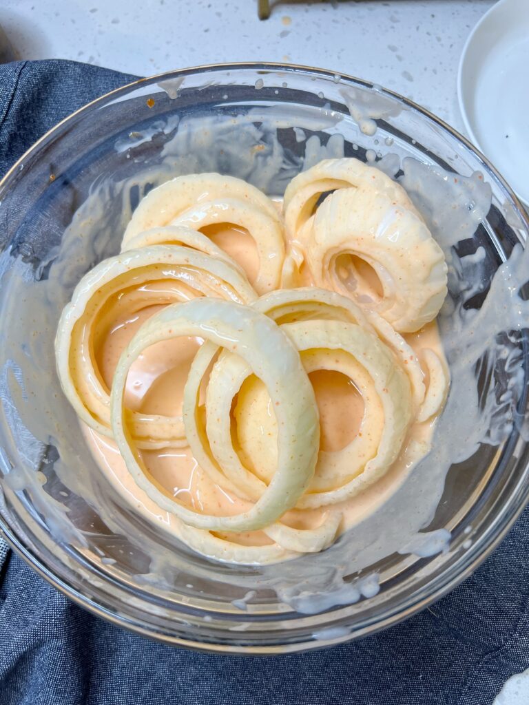 Raw, sliced Vidalia onion rings soaking in a bowl of a buttermilk and Sriracha mixture.