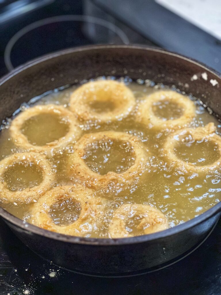 Eight Vidalia onion rings frying in oil in a cast iron pan.