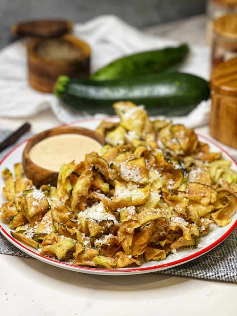 A white, round platter holding crispy fried zucchini ribbons that are sprinkled with freshly grated Parmesan cheese. A small wooden bowl of horseradish sauce is also on the plate.