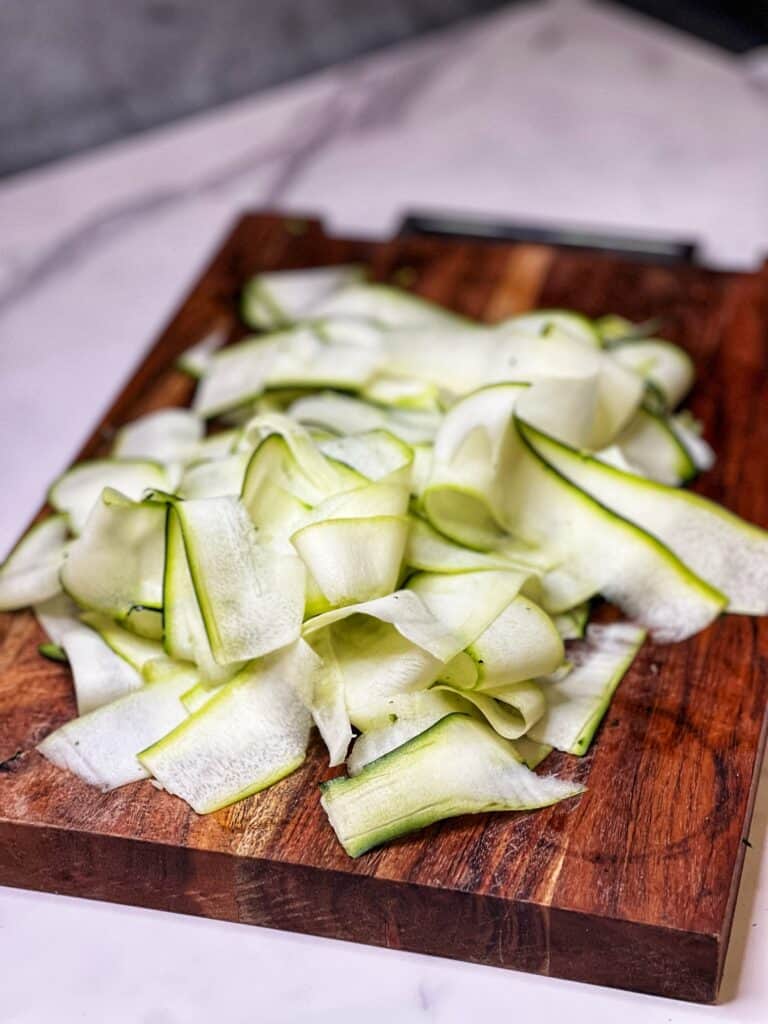 Zucchini that has been thinly sliced into ribbons with a vegetable peeler sitting on a wooden cutting board.