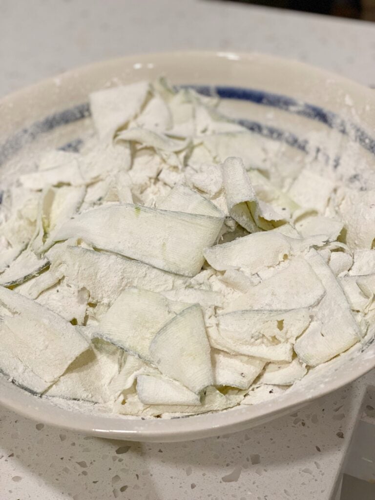 Thinly sliced zucchini coated in flour in a white bowl with blue trim.