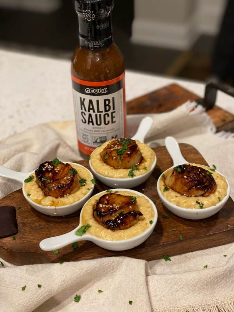 four mini white serving pots filled with corn purée and topped with a Kalbi marinated seared scallop with a bottle of Kalbi sauce in the background