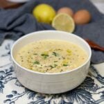 A white bowl filled with Avgolemono Soup. It's a Greek lemony chicken soup. The soup is creamy and is drizzled with olive oil and garnished with fresh parsley and black pepper.