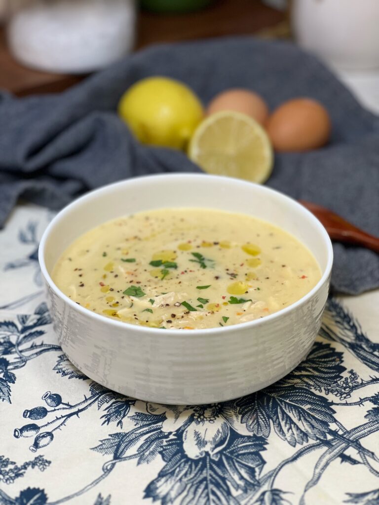 A white bowl filled with Avgolemono Soup. It's a Greek lemony chicken soup. The soup is creamy and is drizzled with olive oil and garnished with fresh parsley and black pepper.