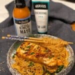 a bowl of spicy sesame noodles topped with a piece of kimchi mayo seared sea bass with a bottle of kimchi mayo and a bottle of gochujang in the background