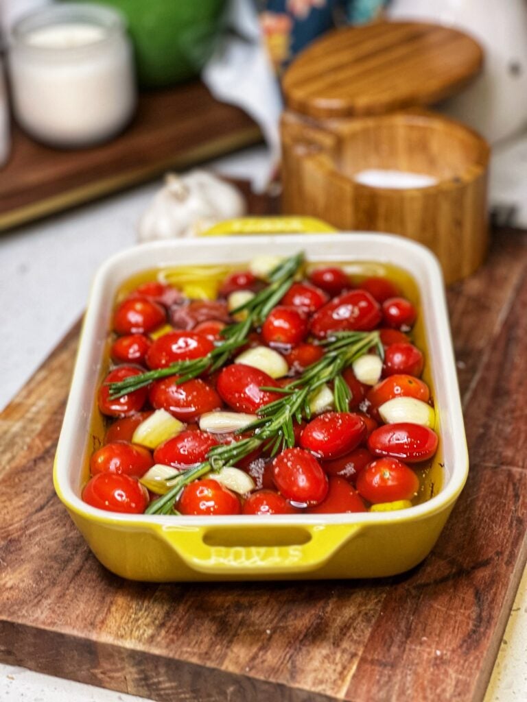 A small, yellow rectangular casserole dish holding cherry tomatoes, garlic cloves and fresh rosemary. Everything is covered in olive oil.