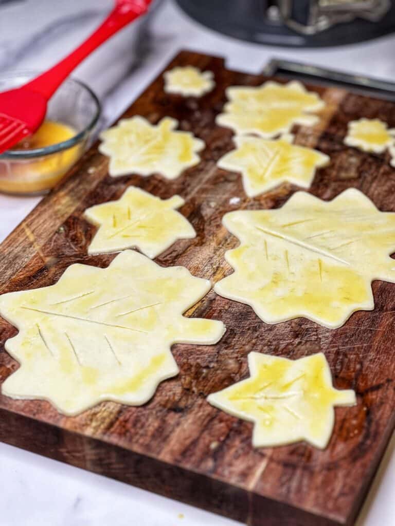 Pastry dough that has been cut into various sizes of leaves sitting on a wooden cutting board.