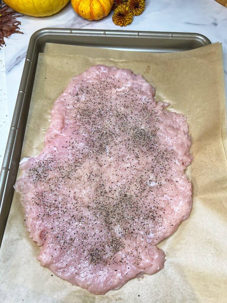 A flattened, boneless turkey breast seasoned with salt and pepper laying on parchment paper inside a sheet tray.