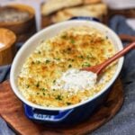 A small blue casserole dish filled with baked, creamy hot clam dip with a wooden spoon in the middle showing what the dip looks like inside. The dip has a top layer of crunchy panko breadcrumbs.