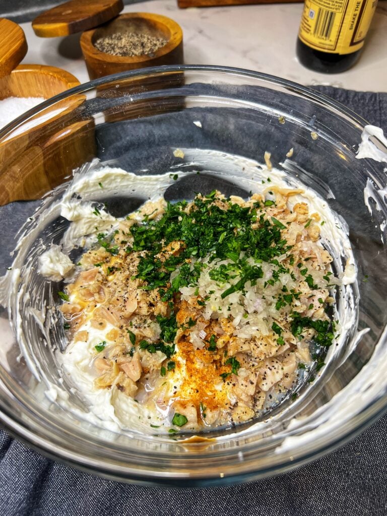 All the ingredients for a creamy hot clam dip in a glass bowl. The ingredients include cream cheese, sour cream, canned clams, spices, and fresh parsley.
