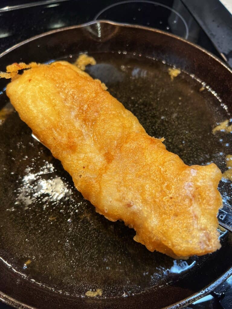 Removing a piece of golden brown, beer battered fish from hot oil in a cast iron pan.