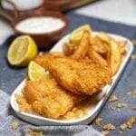 A platter of beer battered fish and chips. The dish is served with lemon wedges with a wooden bowl of homemade tartar sauce in the background.