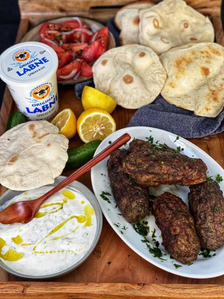 an overhead shot of a wooden platter with a plate of lamb koftka kabobs with labne tzatziki, fresh pitas, lemons and a container of Karoun labne