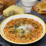 A white bowl with a yellow rim filled with Pasta Fagioli that is garnished with fresh parsley, grated Parmesan cheese and a drizzle of olive oil.
