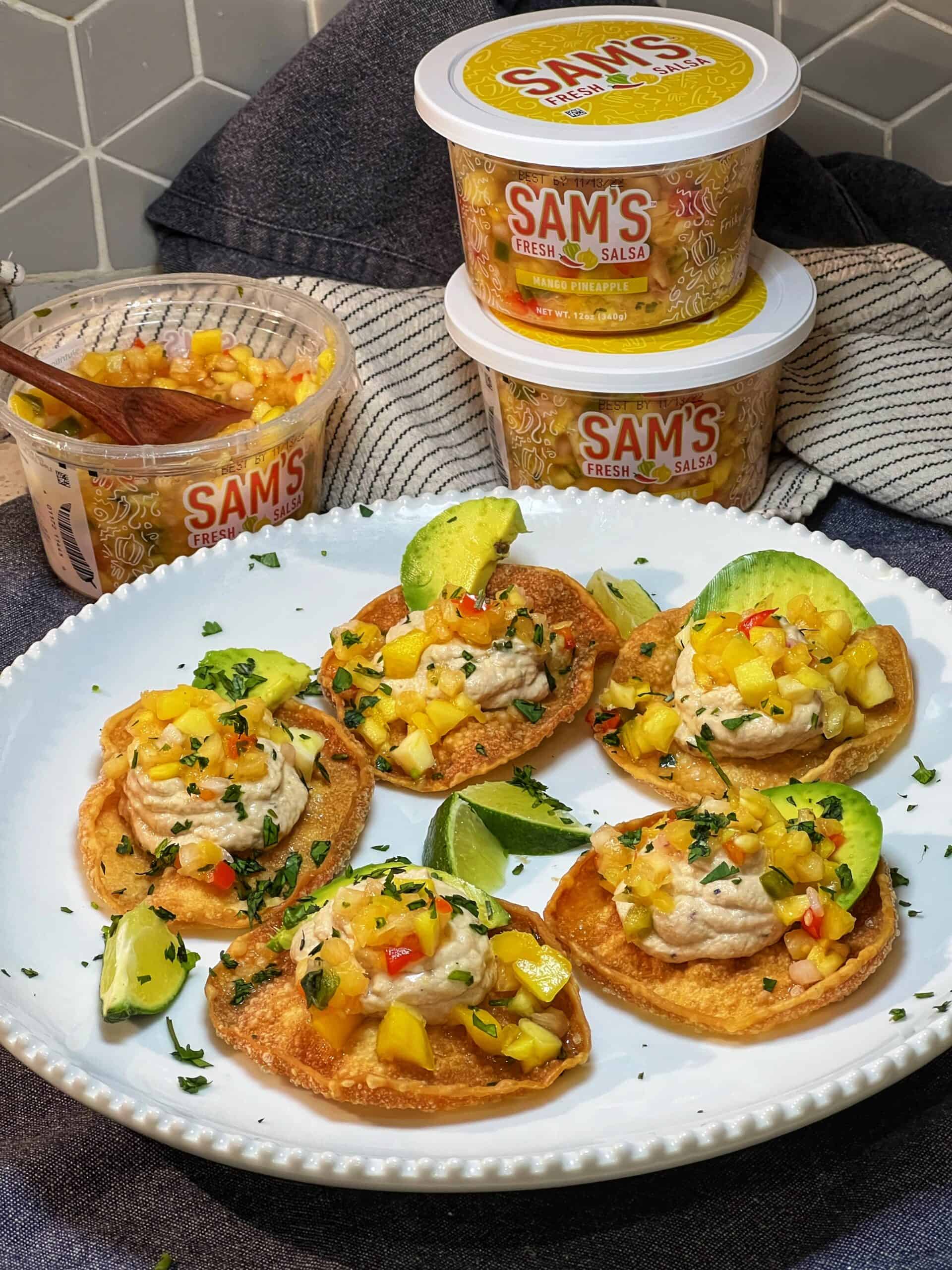 Tuna Mousse on Crispy Fried Wontons with Mango Pineapple Salsa garnished with fresh cilantro and avocado served on a white platter with containers of Sam's Fresh Salsa in the background