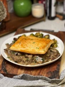 A white shallow bowl of beef tips in cream sauce with mushrooms. The dish is topped with a square of baked puff pastry.