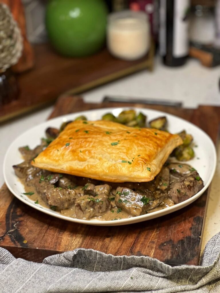 A white shallow bowl of beef tips in cream sauce with mushrooms. The dish is topped with a square of baked puff pastry.
