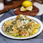 A white platter trimmed in black holding three large pieces of pan fried cod with a lemon butter sauce spooned on top of the fish and garnished with fresh parsley and served with lemon wedges.