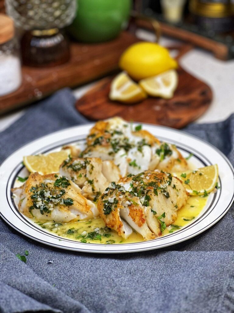 A white platter trimmed in black holding three large pieces of pan fried cod with a lemon butter sauce spooned on top of the fish and garnished with fresh parsley and served with lemon wedges.