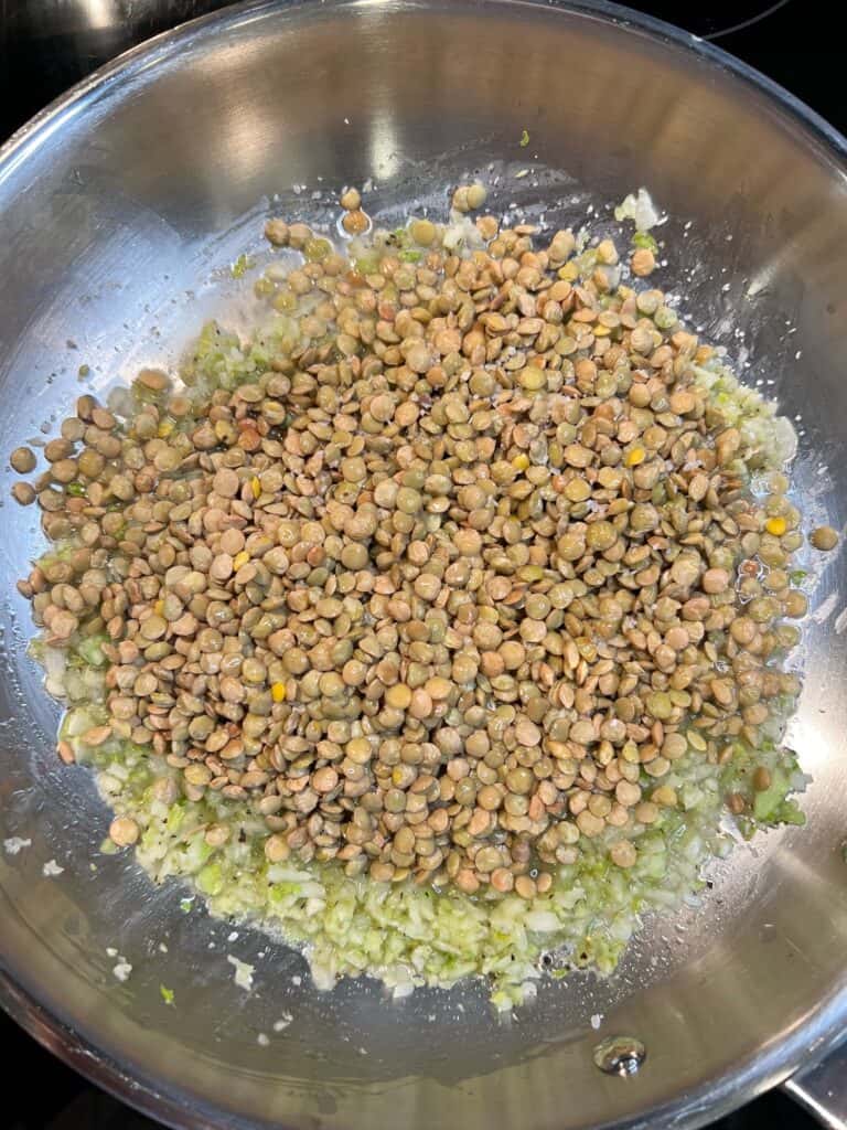 Brownish lentils added to a stainless steel pan of sautéed onions and celery.