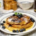 A white plate trimmed in blue holding a stack of three super fluffy blueberry pancakes with syrup, blueberries and mint leaves on top.