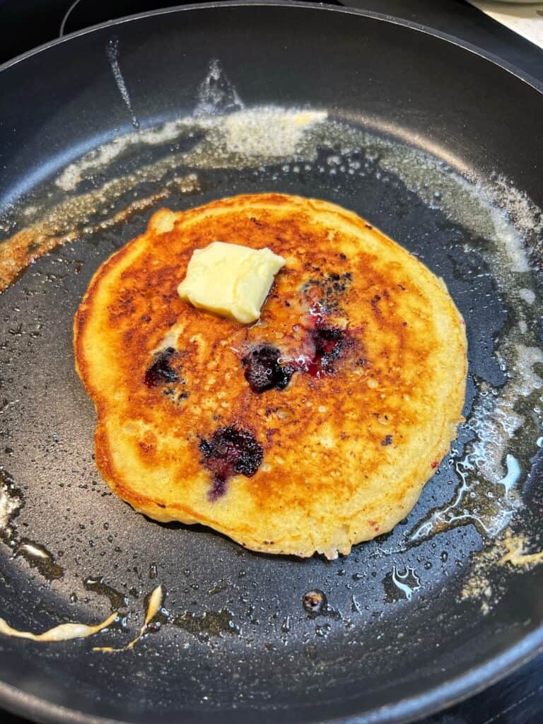 A golden brown, blueberry pancake topped with a pat of butter sitting in a non-stick pan.