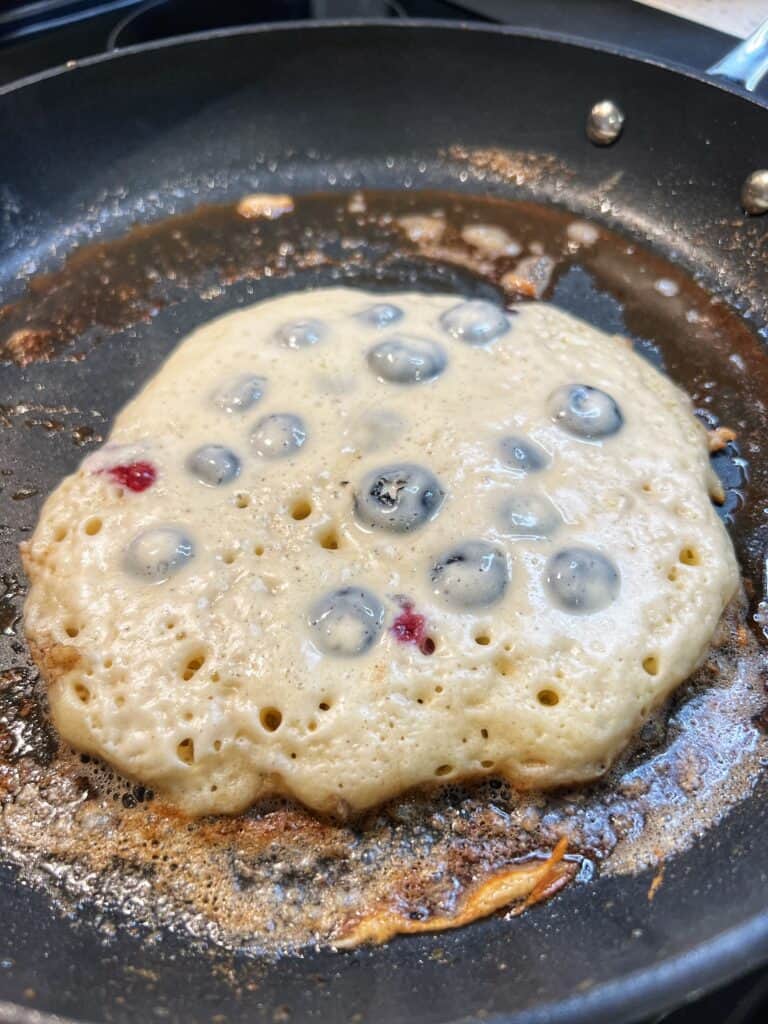 A blueberry pancake with large bubbles on the top and the edges crispy indicating it's ready to be flipped in a non-stick pan.