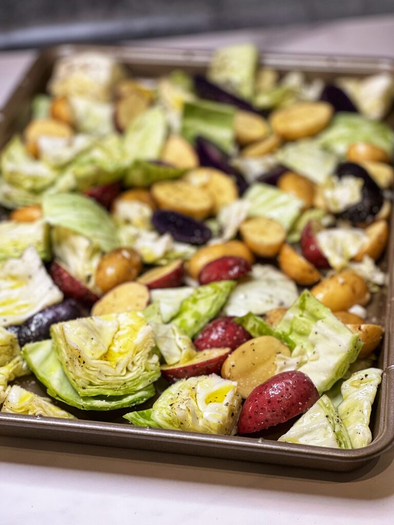 A gold sheet pan filled with large diced cabbage and halved baby potatoes. Both are coated in olive oil and seasoned with salt and pepper.