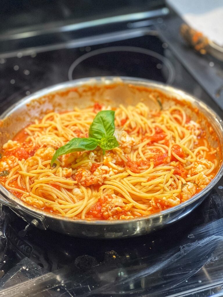 A stainless steel pan holding spicy lobster pasta pomodoro, garnished with fresh basil. The sauce is tomato-based, with chunks of poached lobster.