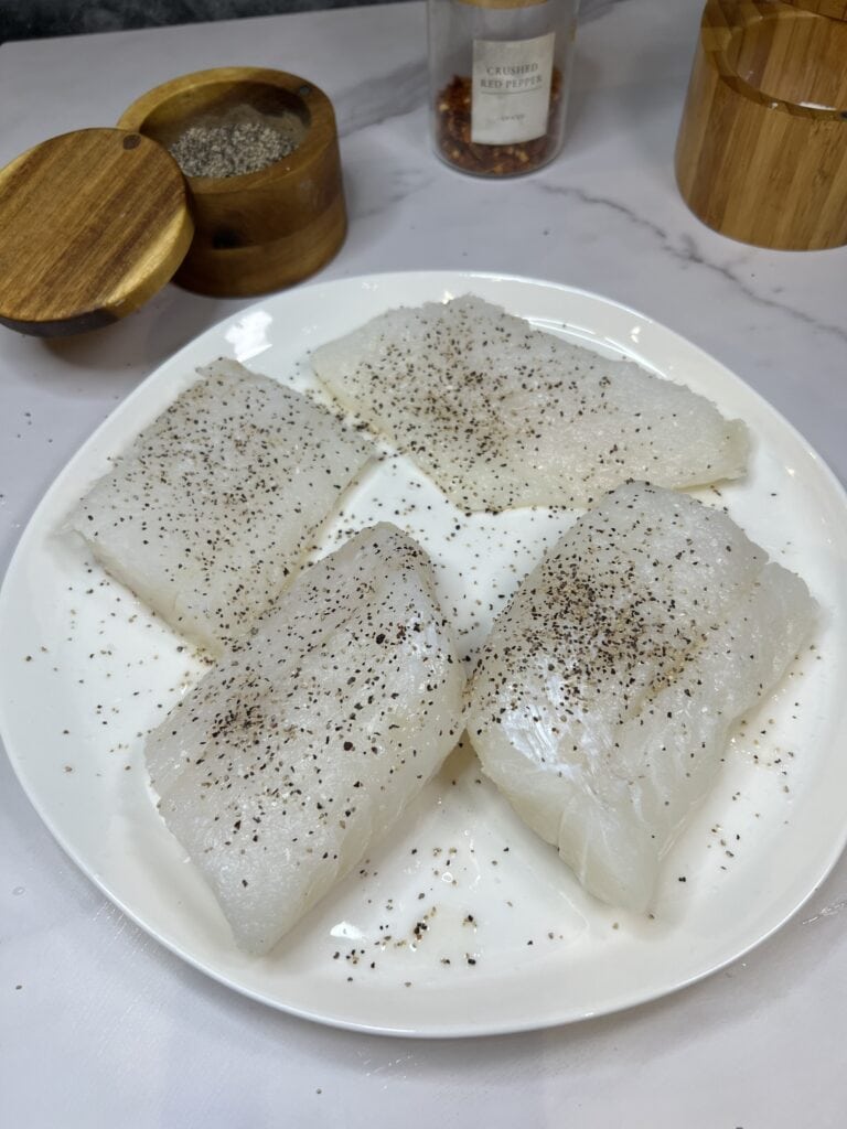 Four cod filets on a white plate seasoned with salt and pepper.
