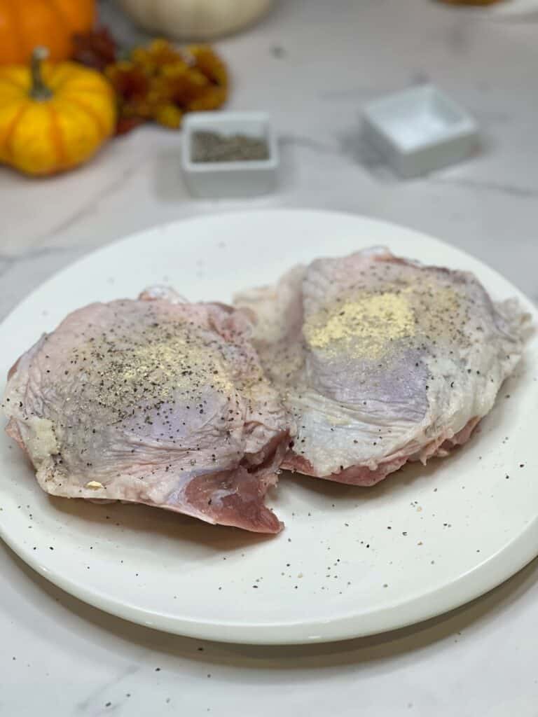 Two bone-in, skin-on turkey thighs seasoned with salt and pepper and laying on a white plate.
