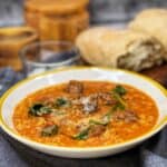 A yellow-rimmed, white bowl filled with Creamy Meatball Soup and garnished with grated cheese with bread in the background.