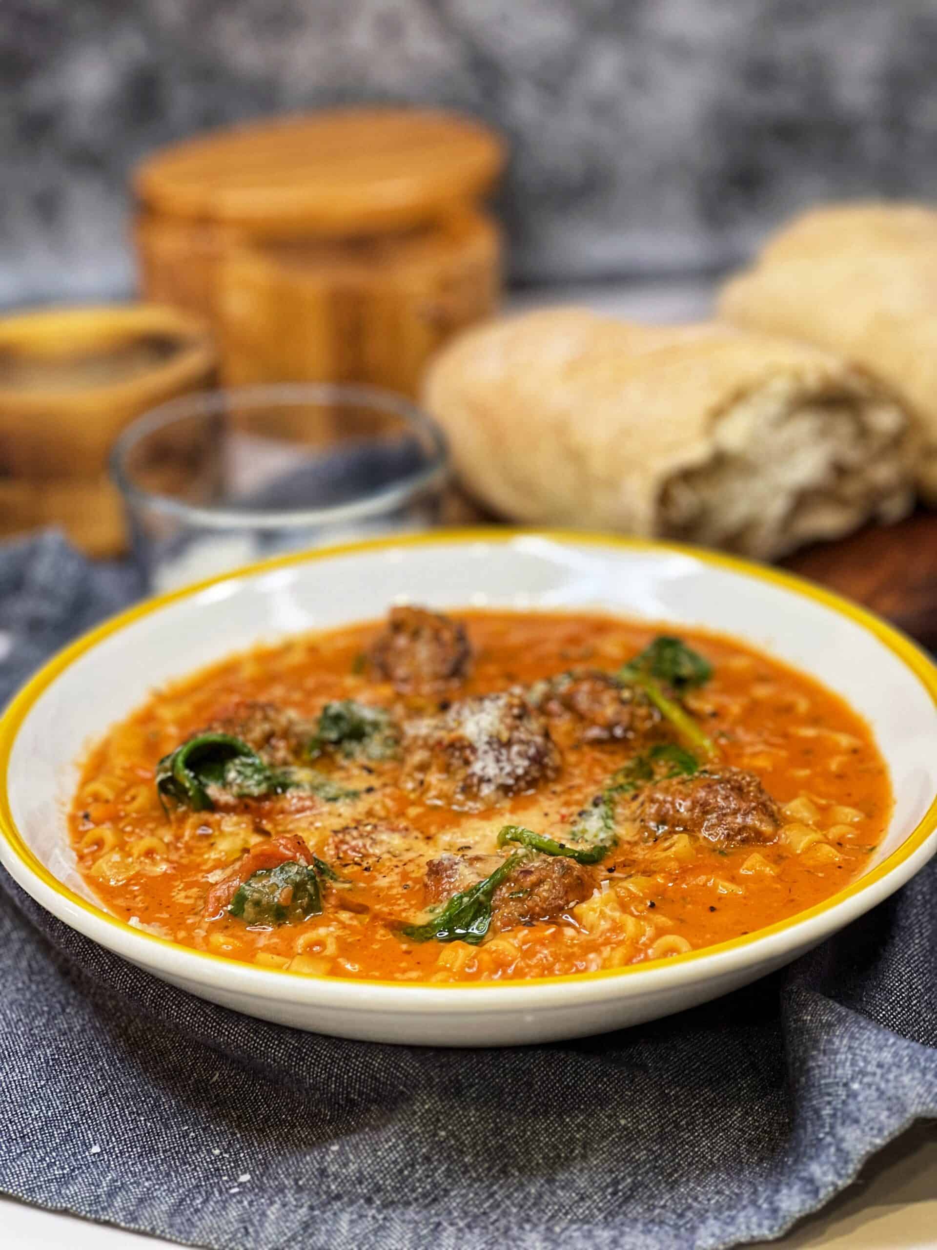 A yellow-rimmed, white bowl filled with Creamy Meatball Soup and garnished with grated cheese with bread in the background.