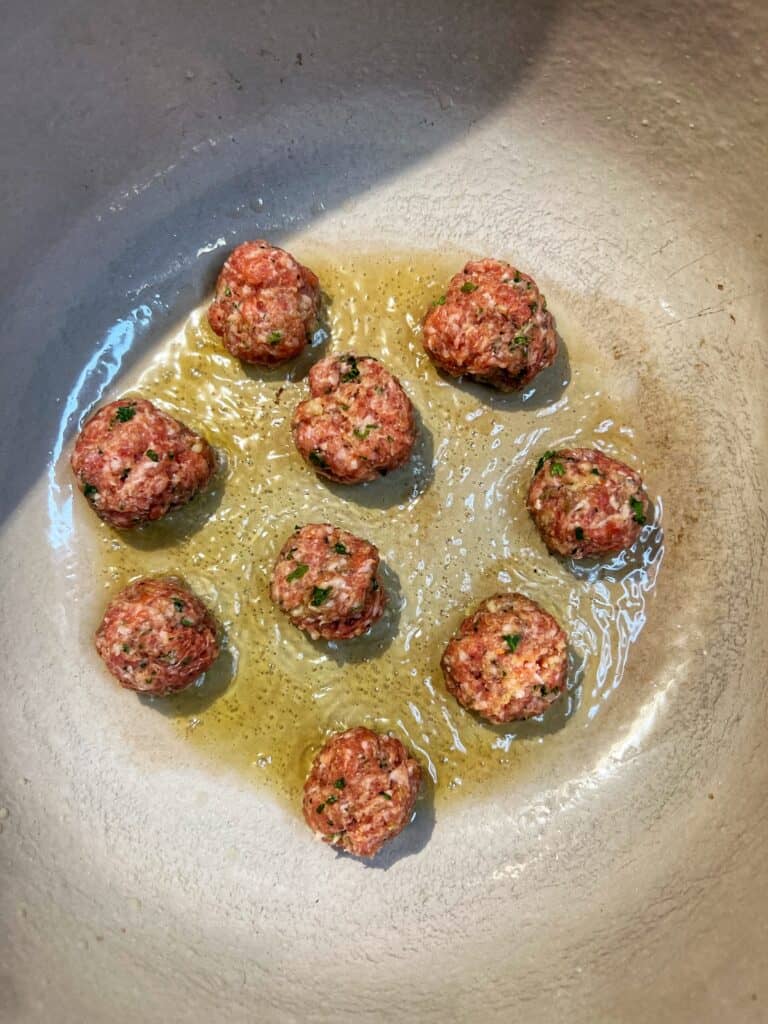 Nine mini meatballs frying in olive oil in a deep pot. 