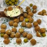 A bunch of fried olives spread across white parchment paper with a brown bowl of garlic aioli dipping sauce.