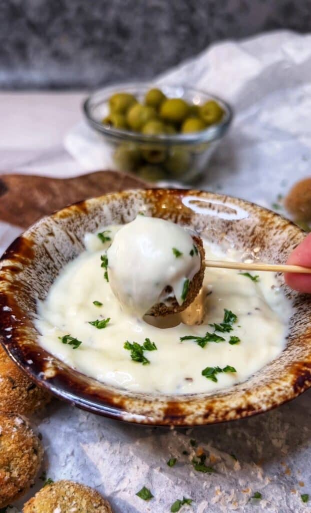Garlic aioli dipping sauce in a brown bowl with a fried olive on a toothpick dipped in the sauce.