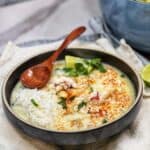 A grey bowl filled with Thai Coconut Soup with Lobster. The soup is garnished with fresh cilantro and a sprinkle of Thai spices. Steamed rice and lemon wedges are also served in the soup.