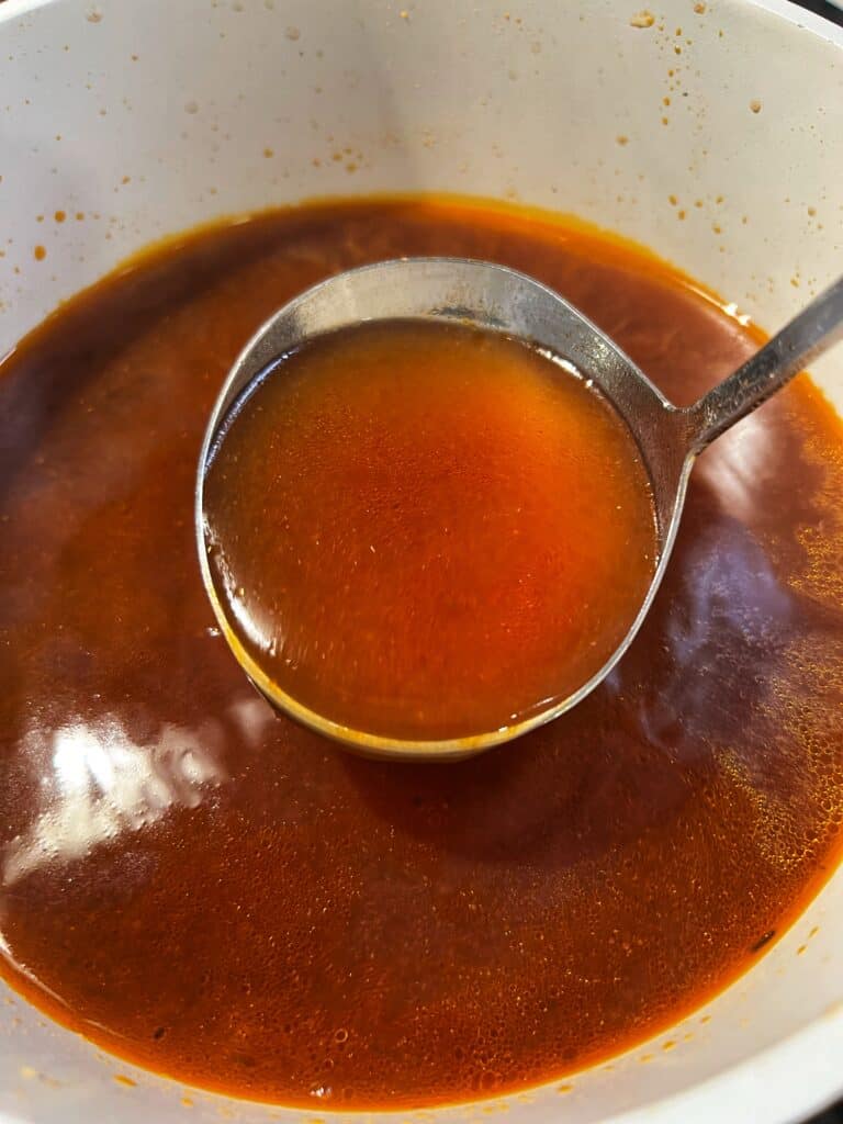 A ladle full of reddish shrimp stock over a pot of shrimp stock.
