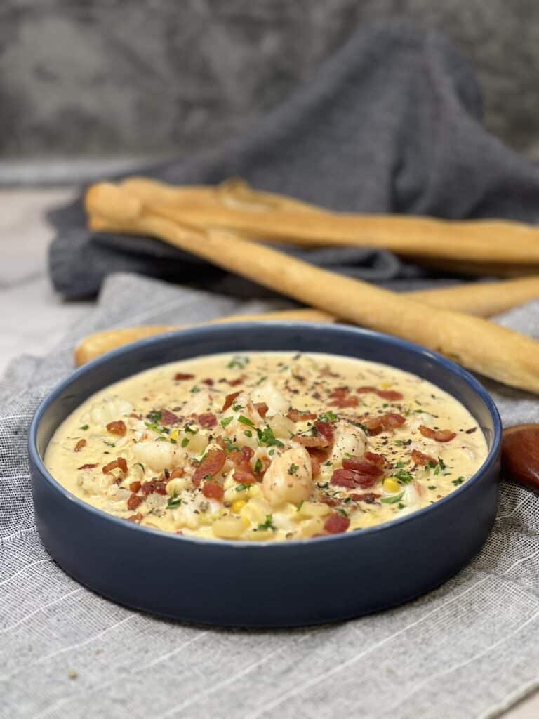 A deep blue bowl filled with homemade shrimp and corn chowder with large breadsticks in the background.
