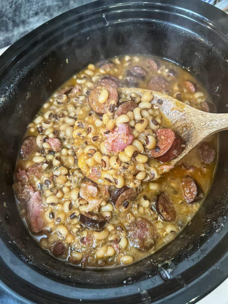 A wooden spoon holding crockpot black-eyed peas with smoked sausage with the crockpot in the background full of the peas. 