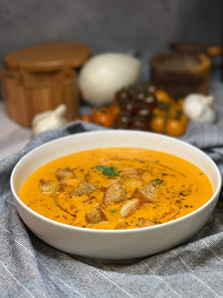 A white bowl filled with roasted tomato basil soup garnished with a drizzle of olive oil, fresh basil, cracked black pepper and homemade croutons.
