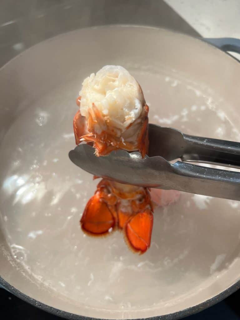 Removing a cooked lobster tail from a pot of simmered water with tongs.