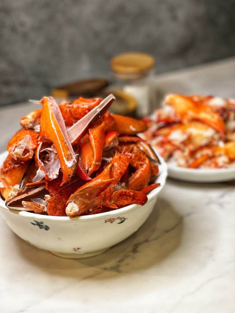 A white bowl of lobster shells with lobster meat on a platter in the background.
